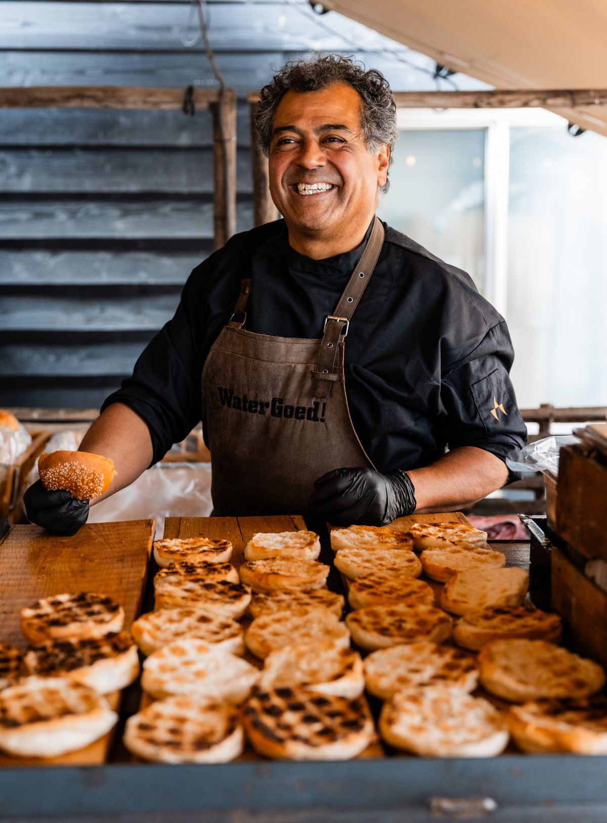 Lachende chef met voor zich een hoop geroosterde hamburgerbroodjes